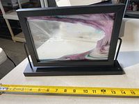 Black framed upright sand art with visible white, purple, and black sand inside, placed on table with tape measure for scale showing about 12 inches width.