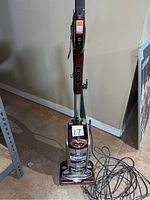Front view of maroon and beige Shark upright vacuum cleaner. Shows power cord uncoiled on floor.