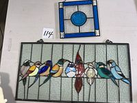 Overhead view of two stained glass pieces lying flat on a table: top small square panel with blue border and central unicorn medallion, bottom larger rectangular panel with colorful birds on clear glass.