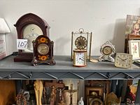 Full view of six clocks arranged on grey shelf showing various styles and sizes including wooden, goldtone, porcelain, and plastic models.