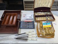 Full overview of lot items on table showing wooden trays, boxed cutlery, steak knives, floral flatware set, and plastic wrapped utensils.