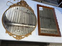 Photo showing both mirrors together on a table, round ornate gold frame with etched mirror on left, rectangular simple wood/gold frame on right