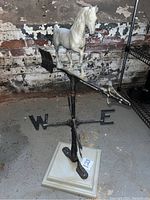 Front view of the weather vane showing white horse figure on arrow pointer with W and E directional markers, mounted on square wooden base.