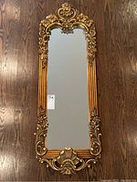Full view of mirror on wood floor showing entire ornate carved gilt frame and mirror surface