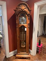 Full front view of floor-standing grandfather clock in oak case with carved bonnet, glass door showing weights and pendulum