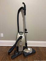 Full front view of Riccar vacuum cleaner with hose, metal wand, and brush head attachment standing upright against the wall.