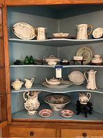 Display of multiple antique porcelain items arranged on cabinet shelves including creamers, sugars, pitchers, plates, and a tureen.