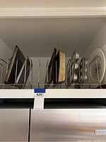 Side view of several metal cookie sheets, baking pans, cupcake pans, and a round plate stored vertically in a wire rack on a shelf.