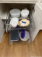 Photo of kitchen cabinet pull-out wire racks showing white Corningware bowls, casserole dish, seashell bowls, and plastic containers with lids.