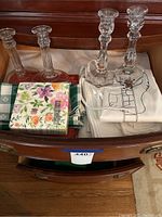 Photo of two pairs of clear glass candlesticks, cloth napkins in green and white checkered pattern, white tablecloth with black graphic design, and multicolored floral paper napkins.