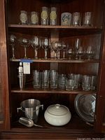 Full view of wooden cabinet shelf holding varied glassware items including branded pint glasses, wine glasses, brandy/snifter glasses, tall drinking glasses, ice bucket, white ceramic pot, metal martini shaker, and tiered glass plates.