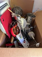 Box containing a variety of kitchen utensils such as red and white cutting boards, metal sieve, whisks, and spatulas.
