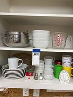 Kitchenware including stainless steel mixing bowls, white porcelain bowls, Pyrex glass measuring cup, assorted mugs, plates, ramekins, salt and pepper shakers, and kitchen timer on shelves.