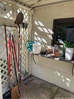 Photo shows an outdoor corner with assorted gardening tools hanging on lattice wall: shovel, rake, broom, garden hoe. A shelf with small terra cotta pots, white plastic pots, and small gardening accessories is visible.