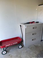 Red Radio Flyer wagon beside three-drawer metal filing cabinet in light gray.