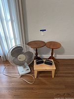 Full lot showing two small round wooden tables, wooden stool, white electric fan, two small black alarm clocks on stool