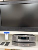 Front view of Dynex TV and Bose Wave Music System main unit shown below it with two remote controls.