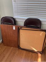 Two foldable card tables with wood finish tops and four brown vinyl padded chairs stacked behind them.