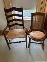 Photo showing two chairs side by side near a window on carpet, one cane rocking chair with cushion and one wooden arm chair with woven seat.