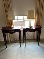 Wide shot showing two identical dark wood hall tables each topped with a table lamp, set in front of a window with beige curtains.