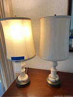 Two table lamps with white marble bases and off-white silk shades seen side by side on a wooden surface.