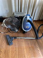 Photo showing Oreck vacuum cleaner, metal fan, and paper shredder on wooden floor in front of curtains.