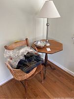 Wide view of vintage octagon-shaped wood table, metal lamp with beige shade, metal bowl on table, coated basket on damaged wooden chair with two dry-cleaned throws inside.