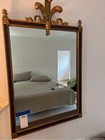 Full front view of the mirror mounted on a wall, showing the rectangular reflective surface and the gold and dark brown painted wooden frame with decorative top piece.