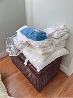 Brown wicker trunk with a stack of white linens including sheets and towels on top, with a blue towel folded on top of the white linens.