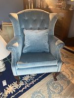 Front view of the blue upholstered wingback armchair showing tufted backrest, rolled arms, matching throw pillow, and wooden legs.
