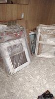 Multiple white picture mats and wooden stitchery frames wrapped in plastic, resting on floor against wood panel wall.