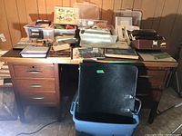 View of wooden desk covered with plastic bins, boxes, papers, art supplies, and a black portfolio case resting on a plastic tote beneath the desk