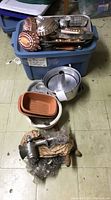 Blue plastic tote filled with vintage and newer baking pans and molds stacked on top; below tote are several pans including a cake ring pan, a small loaf pan, and a plastic carrier for molds; hand crank meat grinder with accessories shrink-wrapped, placed on floor.