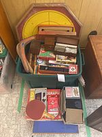 Overview of lot showing a large green bin filled with multiple wooden game boards, boxed cards, large wooden dice, a yellow ring toss target, and scattered game components on the floor in front of the bin.