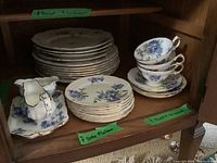 Full shelf with stacked dinner plates, side plates, saucers, three stacked teacups, and a creamer on a matching tray, all with floral blue-purple pattern and gold trim.