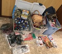 Overview of multiple clear and blue plastic storage totes filled with assorted Christmas decorations including glass ball ornaments in red, blue, silver; pine cones; ribbons; and packing materials on the floor next to a wall.
