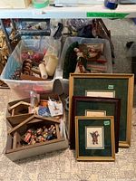 Wide shot showing two large totes filled with Christmas decorations including ornaments, candles, cards, and boxed nativity sets, along with three framed Santa-themed cross stitch art pieces leaning against the totes.