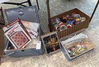 Sewing basket with quilting books, finished embroidery projects, and assorted needlework materials; box of curling ribbons rolled and stored nearby; lidded plastic tote with additional craft supplies visible.