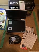 Photo of ONN portable DVD player open with screen up, Lumix digital camera, instruction papers, and Sandisk cards on carpet.