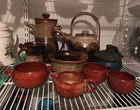 Full view of the collection of glazed pottery items including the tall coffee pot, tea pot, jams jar, bowl, and various cups and jug, displayed on a wire shelf.