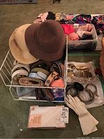 Image showing two hats on top of a white crate filled with belts and a nearby box with gloves and costume jewelry on a carpeted floor.