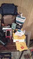 Photo showing camera bags, boxed Argus slide viewer, camera lens boxes, and miscellaneous photography accessories stacked on a table.