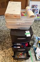 Photo showing two snap-lid boxes stacked on top of a black three-drawer plastic storage unit with two drawers open containing stationery and craft supplies.