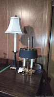 Two table lamps side by side on a wooden surface against wood panel wall. Taller lamp has black ceramic and metal base with white taper shade; shorter lamp is gold tone with two bulbs and black shade.