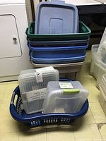Stack of six plastic storage totes in green, blue, beige with matching lids, clear plastic containers in a blue laundry basket on floor near laundry machines.