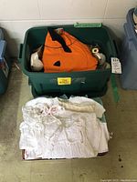 Photo showing green tote bin with orange pumpkin costume inside, white folded vintage baby clothes at front.