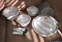 Full set of four white ceramic casseroles with glass lids, two detachable handles, and two plastic storage lids displayed on a beige surface