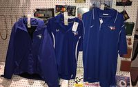 Three Blue Jays branded apparel items hanging on pegs: jacket, Under Armour long sleeve, and Nike polo shirt.