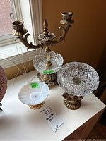 Photo of an ornate metal candelabra with two candle holders with three pressed glass bowls with metal bases on a table near a window.