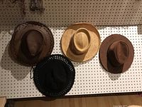 Four hats hanging on pegboard: brown leather outback hat, natural straw Panama Jack hat, brown suede or leather hat, black crochet style ladies hat.